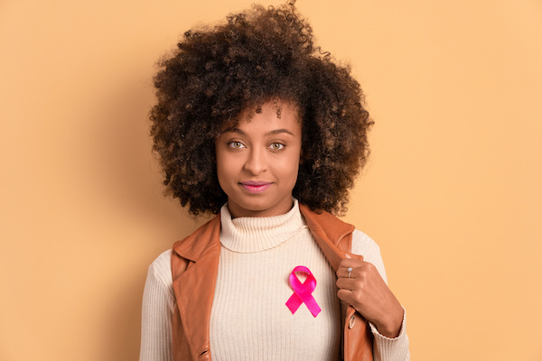 Woman in white sweater pulls back brown vest to reveal pink awareness ribbon