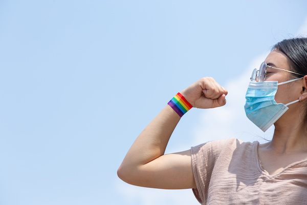Woman with rainbow wristband and face mask flexes bicep over blue background