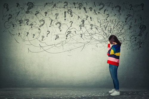 woman in multi-colored sweater holding her hand to her forehead against a grey background covered with question marks