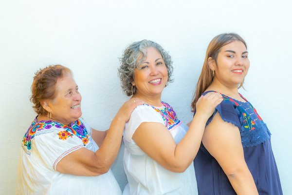 Three generations of smiling Mexican women with blouses with floral patterns in a row holding their shoulders against a white background