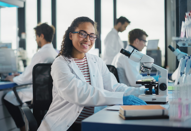 Young black woman in white lab coat is sitting at a lab table with a microscope, smiling at the camera