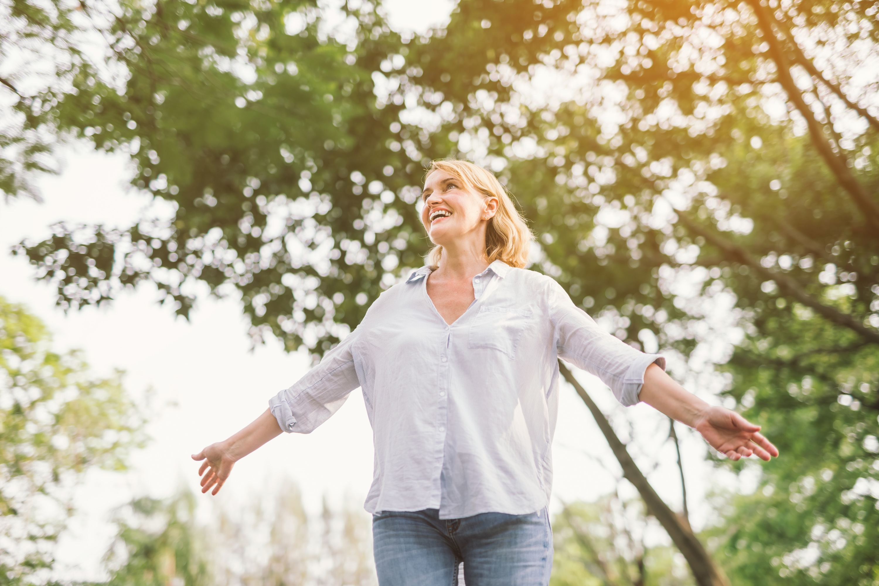 Woman with blonde hair standing under tree with arms open wide