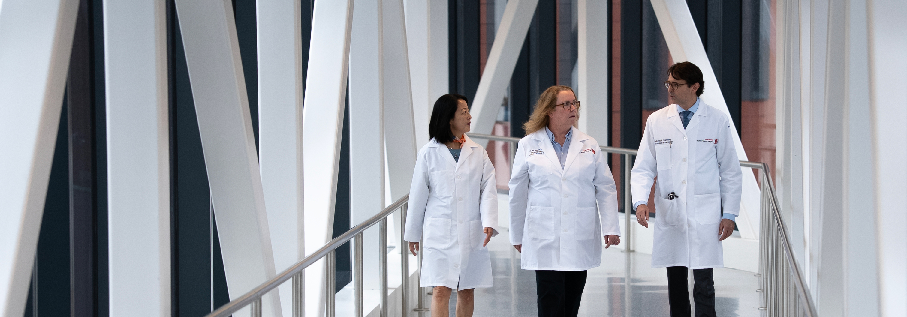 Three researchers in white lab coats talking while walking in skybridge hallway